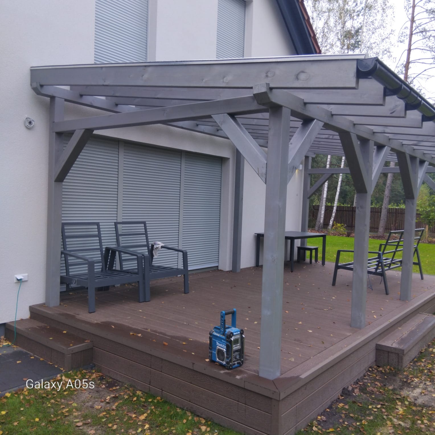 Zadaszony taras z drewnianą podłogą i szarymi meblami ogrodowymi, widok na dom z roletami zewnętrznymi i zielony trawnik w tle.