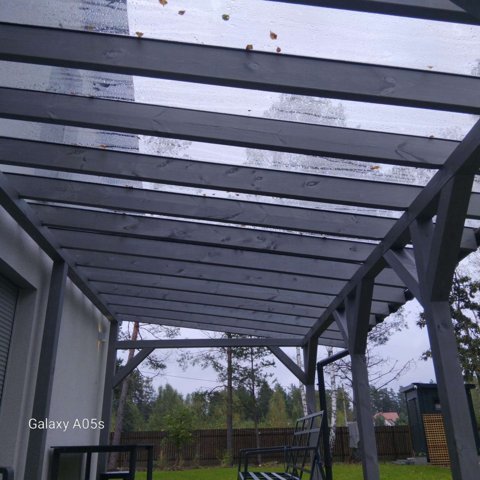 Drewniana pergola pomalowana na szaro, z przezroczystym zadaszeniem, widok z dołu na tle zieleni i domu. Widoczne krople deszczu na zadaszeniu.