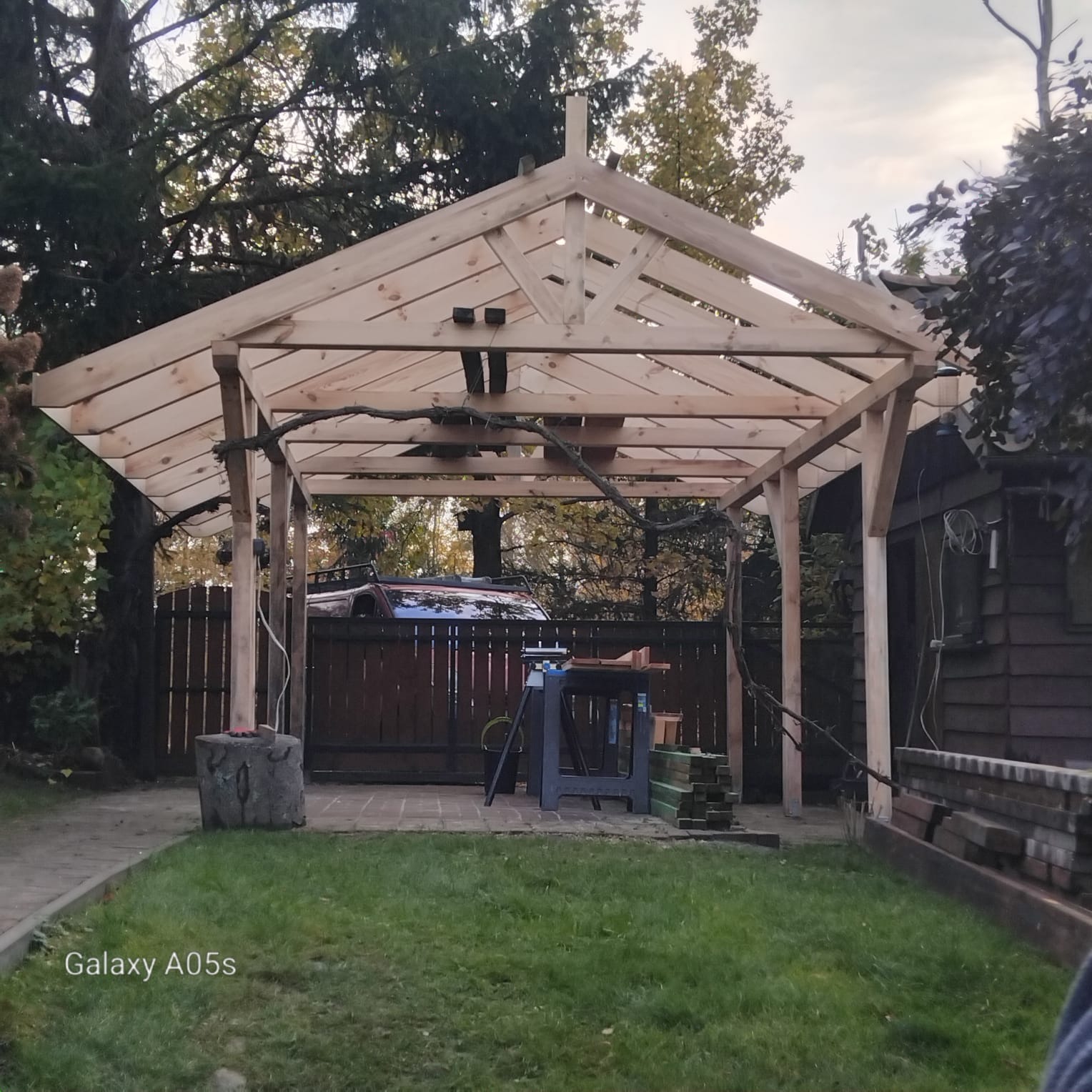 Drewniana konstrukcja altany w trakcie budowy, widoczne narzędzia i materiały budowlane na trawniku, tło z zielenią drzew i zabudowaniami.