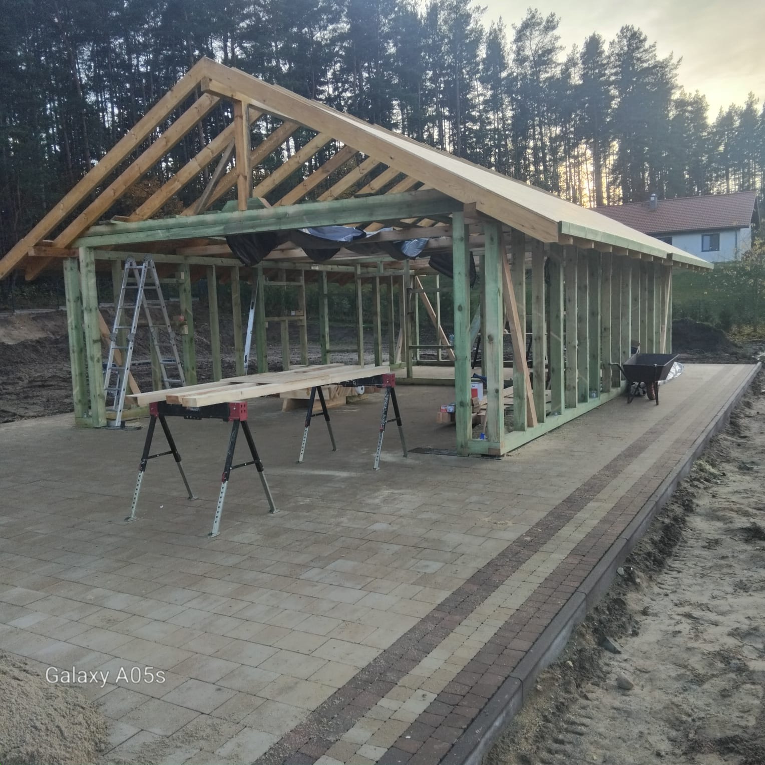 Konstrukcja drewniana zadaszenia w trakcie budowy, widoczne elementy więźby dachowej i ściany szkieletowe. W tle las i dom, na pierwszym planie kostka brukowa.