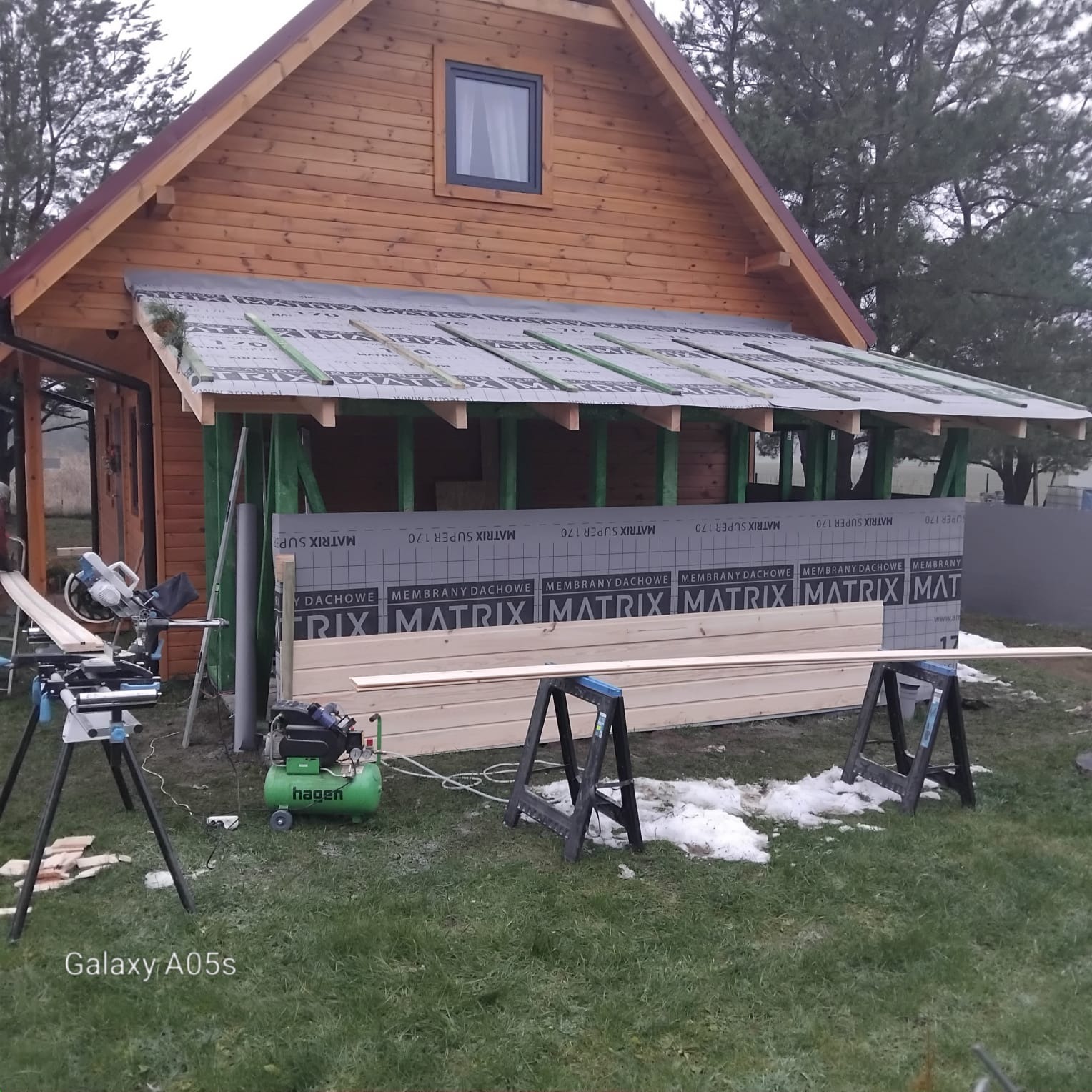 Drewniany dom w trakcie prac elewacyjnych: widoczne membrany dachowe, deski, narzędzia (piła, kompresor). Częściowo ośnieżony trawnik.