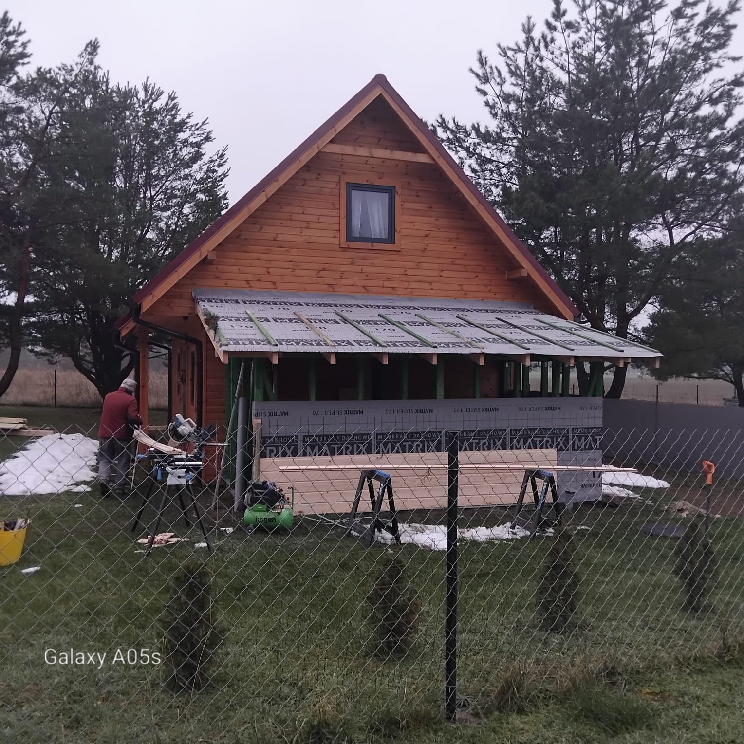 Budowa drewnianego domku z dobudowanym zadaszeniem. Widoczny pracownik przy pracy, narzędzia i materiały budowlane. Częściowo ośnieżony teren.