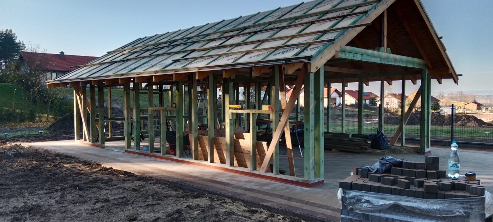 Konstrukcja z drewna w Dywitach, zadaszenie w trakcie budowy. Widoczne elementy konstrukcyjne i materiały budowlane. Prace w toku, teren przygotowany pod dalsze etapy.