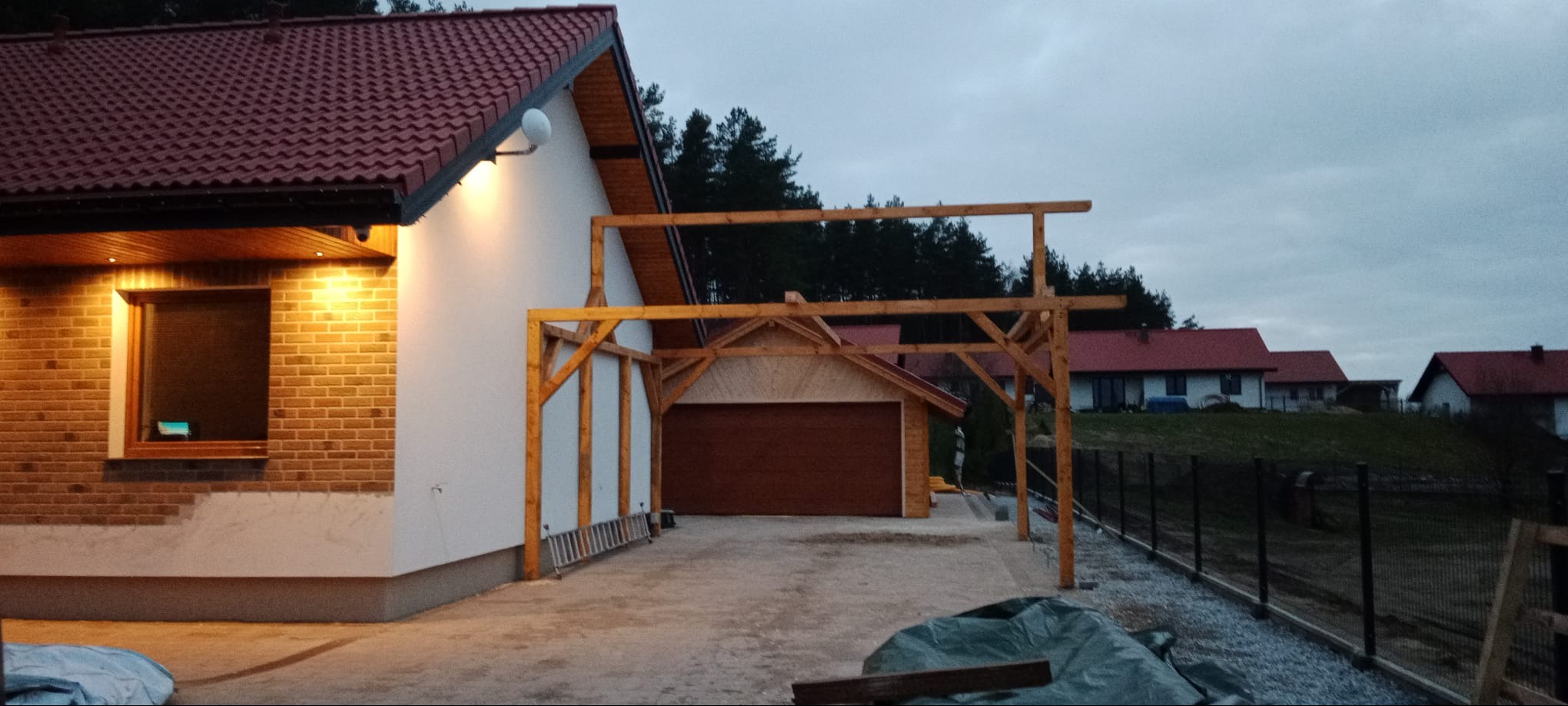 Nowoczesny dom z ceglaną elewacją i drewnianą konstrukcją wiaty garażowej w trakcie budowy, widok z zewnątrz, wieczorne oświetlenie.