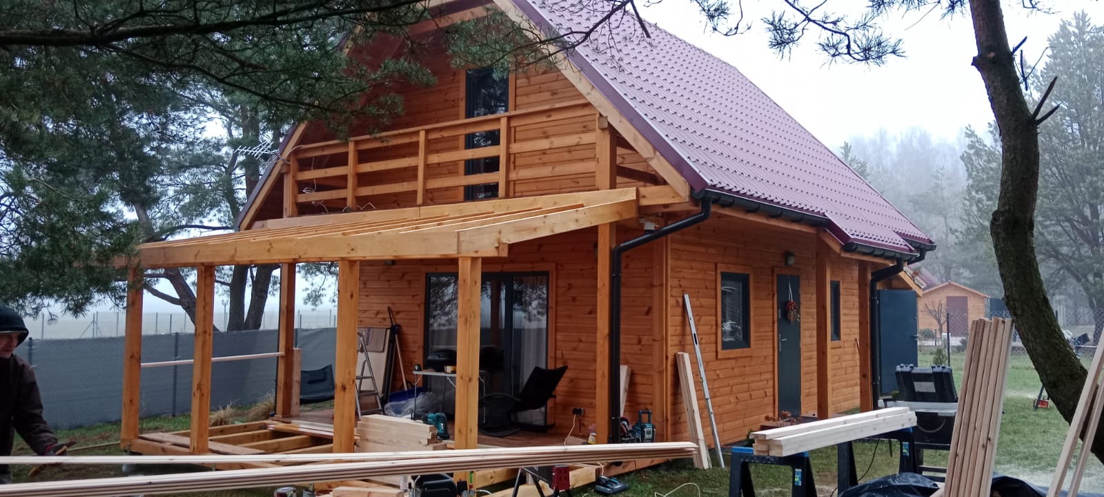Drewniany dom z czerwonym dachem w trakcie budowy, widoczne elementy konstrukcyjne werandy i narzędzia. Tło stanowią drzewa i mglista pogoda.