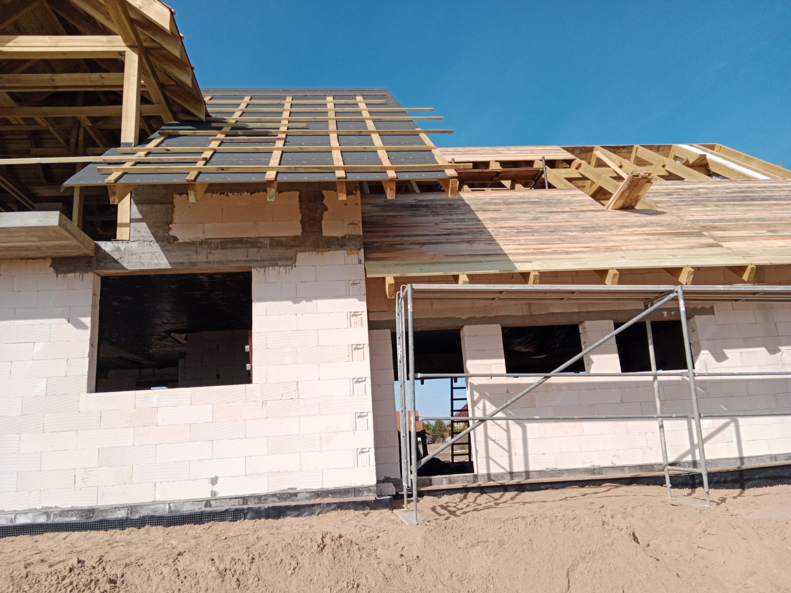 Budowa domu w trakcie: ściany z białego bloczka, częściowo zadaszony drewnianą konstrukcją dachu, widoczne rusztowania i piasek. Błękitne niebo w tle.