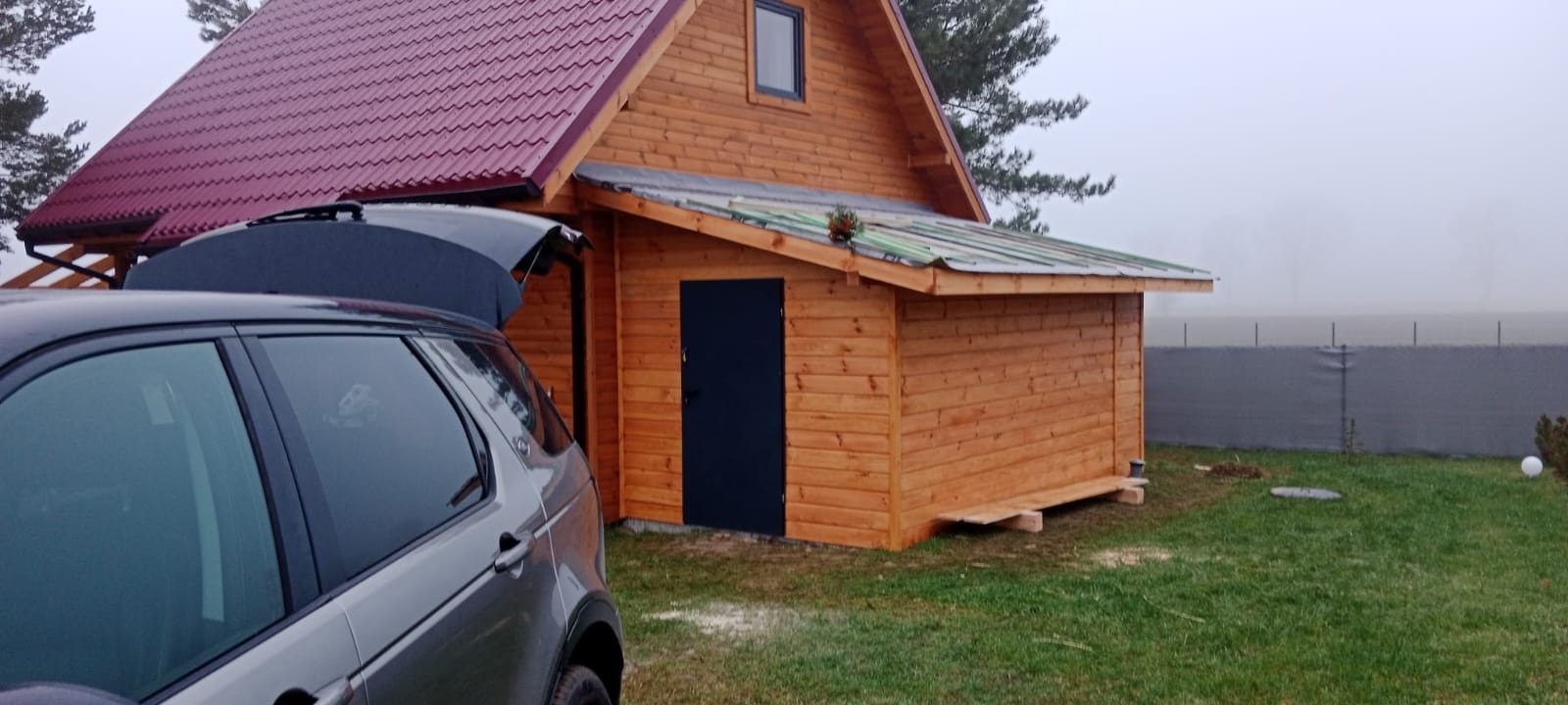 Drewniany domek z ciemnymi drzwiami i czerwonym dachem, częściowo zasłonięty przez szary samochód z otwartym bagażnikiem na tle mglistego krajobrazu.