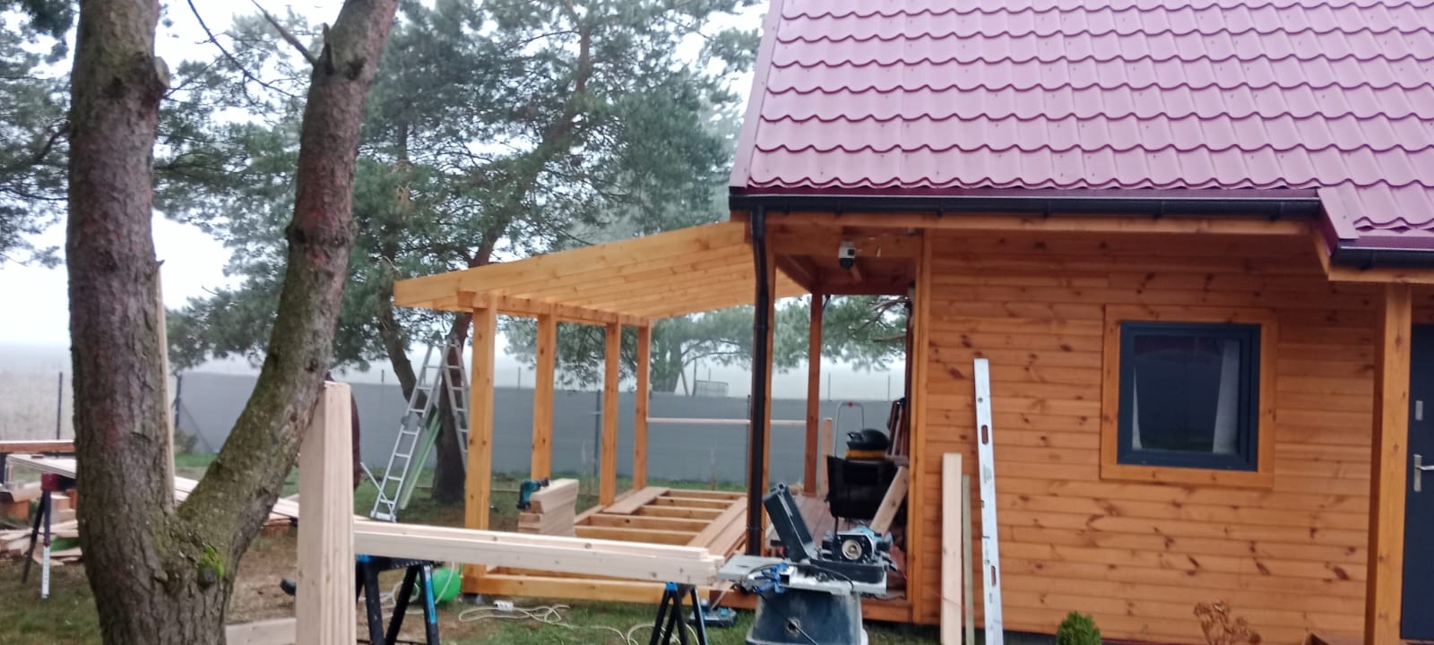 Budowa drewnianej altany przy domu z czerwonym dachem. Widoczne narzędzia i materiały budowlane. Prace w toku, konstrukcja w surowym drewnie.