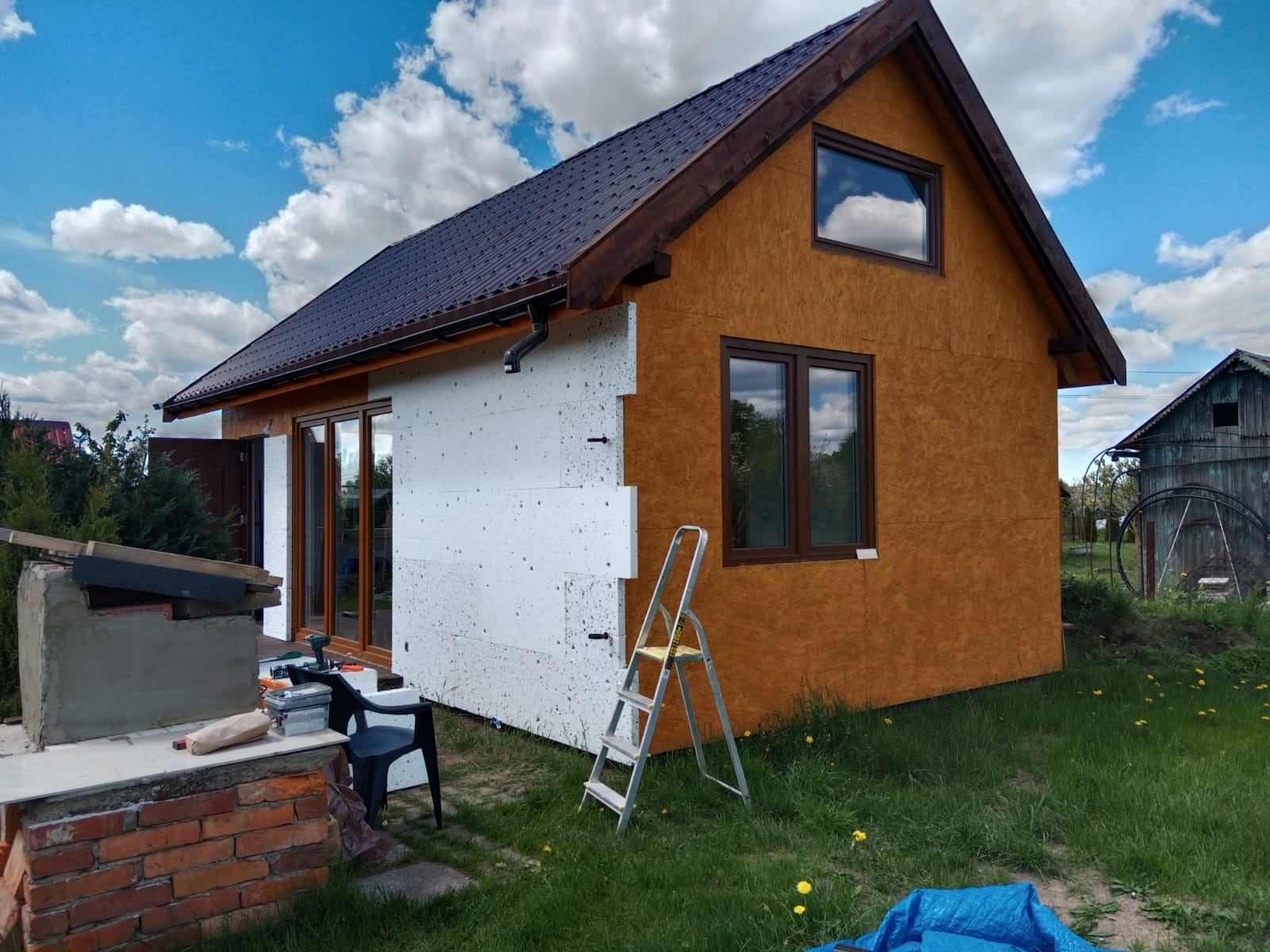 Dom w trakcie ocieplania: biały styropian na jednej ścianie kontrastuje z surową płytą OSB na pozostałych. Drabina oparta o ścianę, narzędzia obok. Błękitne niebo z chmurami w tle.