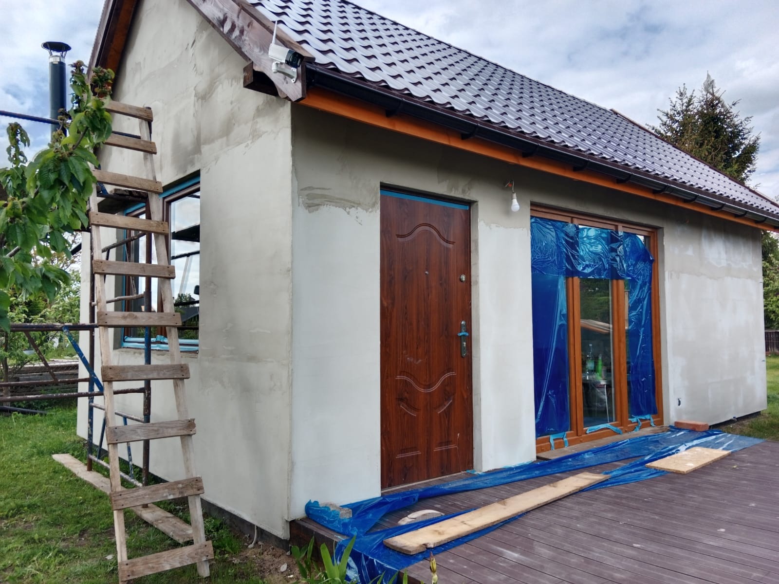 Dom w trakcie prac elewacyjnych: szpachlowanie ścian zewnętrznych, zabezpieczone okna folią, drewniana drabina oparta o ścianę. Widoczny fragment drewnianego tarasu.