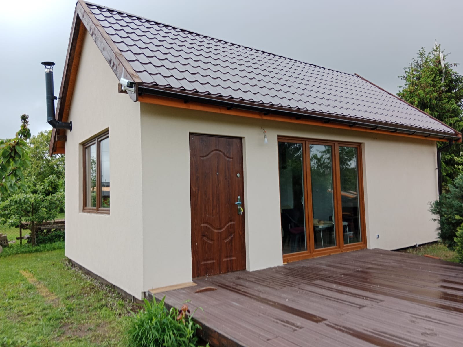 Otynkowany, parterowy budynek z brązowym dachem, drewnianą stolarką okienną i drzwiami. Widoczny fragment drewnianego tarasu i zieleni. Elewacja w jasnym odcieniu beżu.