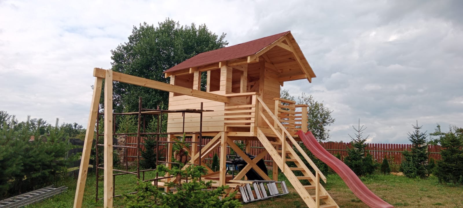 Drewniany plac zabaw z domkiem, schodami i zjeżdżalnią, w trakcie budowy. Widoczne rusztowanie i drabina. Czerwony dach domku na tle zieleni.