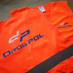 Dwa pomarańczowe bezrękawniki ostrzegawcze z nadrukowanym logo 'DRÓG POL' w kolorze białym i niebieskim oraz szarym pasem odblaskowym, leżące na drewnianej powierzchni.