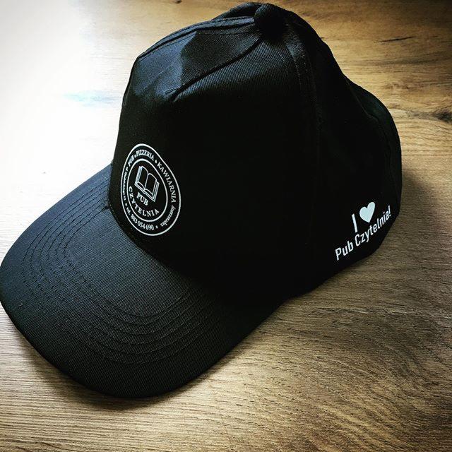 Czarna czapka z daszkiem, z nadrukowanym logo 'Pub Czytelnia' w formie okrągłego znaku graficznego z książką w środku, oraz napisem 'I love Pub Czytelnia!' z boku, leżąca na drewnianym blacie.