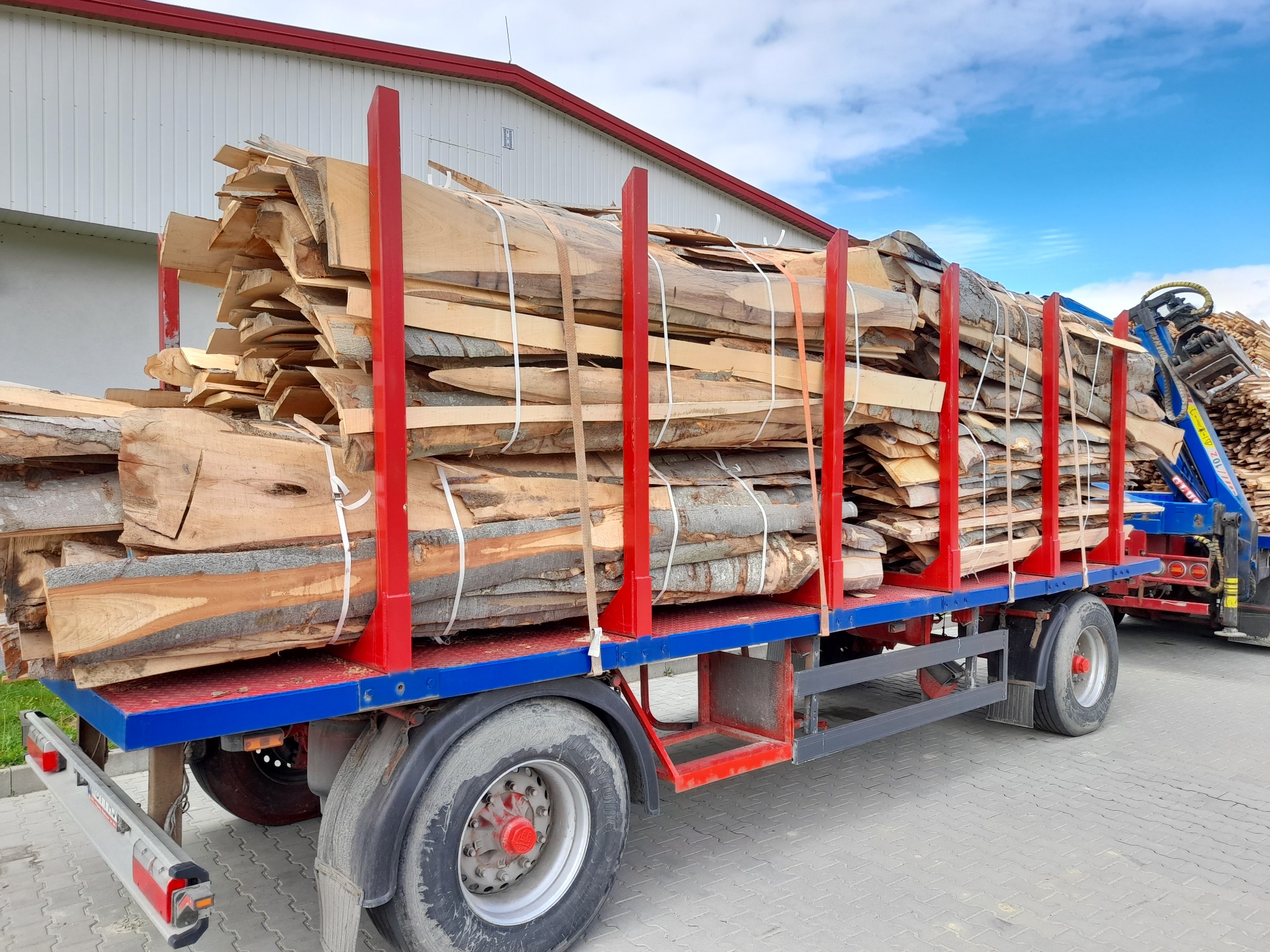 Ciężarówka z czerwoną ramą i niebieską platformą załadowana pociętym drewnem kominkowym, zabezpieczonym białymi pasami, na tle magazynu z czerwonym dachem i błękitnego nieba.