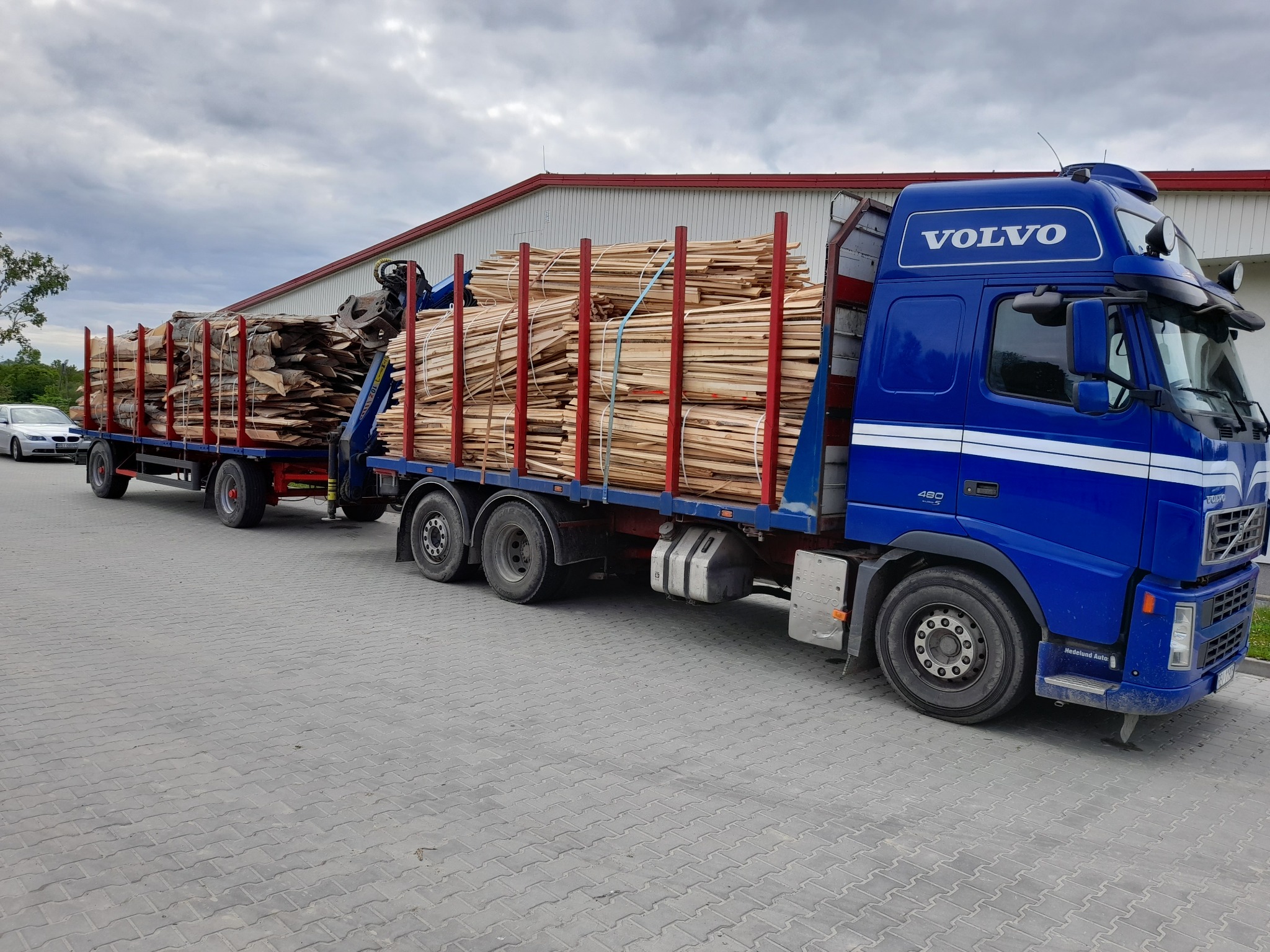 Ciężarówka Volvo z przyczepą załadowana pociętymi kawałkami drewna, zabezpieczonymi pasami, na tle szarej kostki brukowej i budynku z czerwonym dachem.