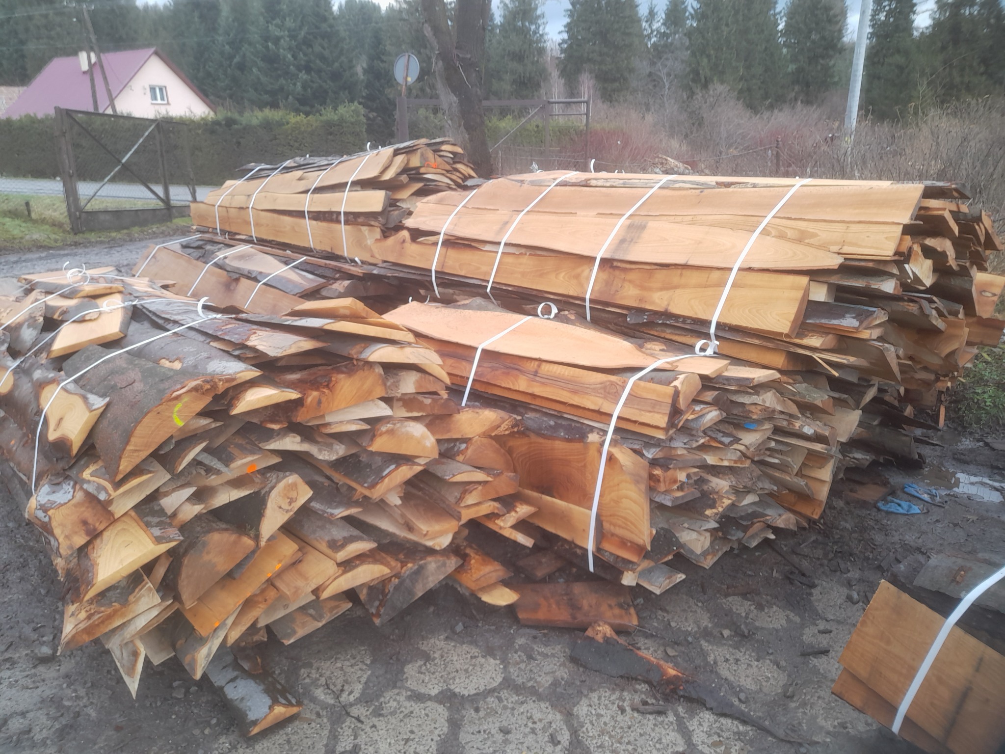 Dwa stosy świeżo pociętych desek z korą, spięte białym sznurkiem, na szarym, prawdopodobnie betonowym podłożu, w tle drzewa i dom z czerwonym dachem.