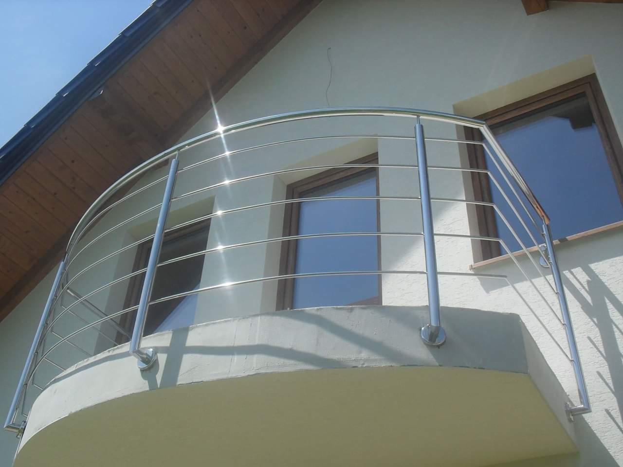 Balkon z zaokrągloną, chromowaną balustradą ze stali nierdzewnej, zamontowaną na betonowej płycie, z widocznymi oknami budynku w tle.