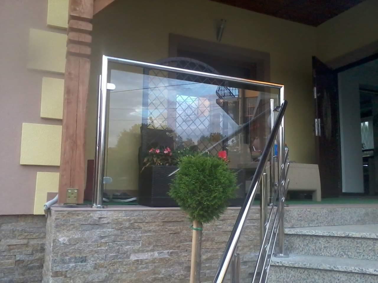 Szklana balustrada ze stalowymi elementami na kamiennym podeście przed wejściem do domu, widoczny mały krzew w donicy.