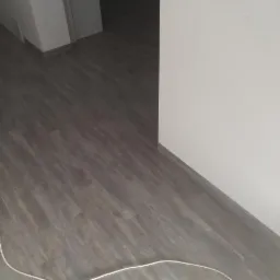 Ułożona, szara wykładzina imitująca drewno w korytarzu, widoczny kabel przy podłodze.