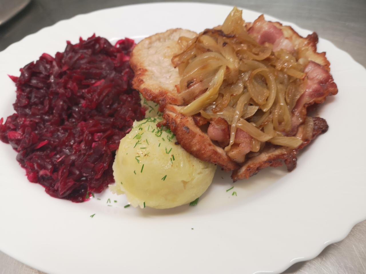 Kotlet schabowy z duszoną cebulą i boczkiem, buraczki i puree ziemniaczane posypane koperkiem, podane na białym talerzu.