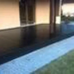 Ciemna, gładka powierzchnia przydomowego tarasu z niskim obrzeżem z jasnych kamieni, widok z poziomu trawnika.