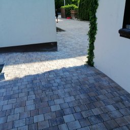 Inkognito - Ułożona kostka brukowa w odcieniach szarości i beżu, przylegająca do białej ściany z pnącą rośliną, widok z perspektywy niskiego kąta.