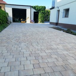 Inkognito - Ułożona beżowo-szara kostka brukowa na podjeździe, widoczna ubijarka w tle, budynek z garażem i fragment trawnika.