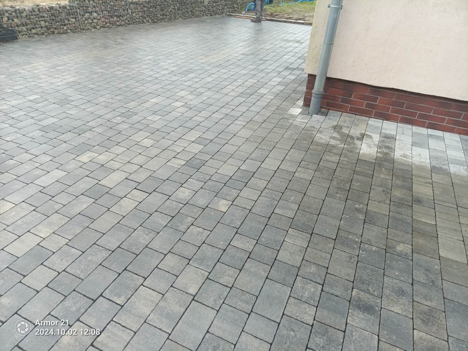 Ułożona szara kostka brukowa na posesji, widoczny fragment elewacji budynku z rynną i obrzeże z kamieni w metalowej siatce.