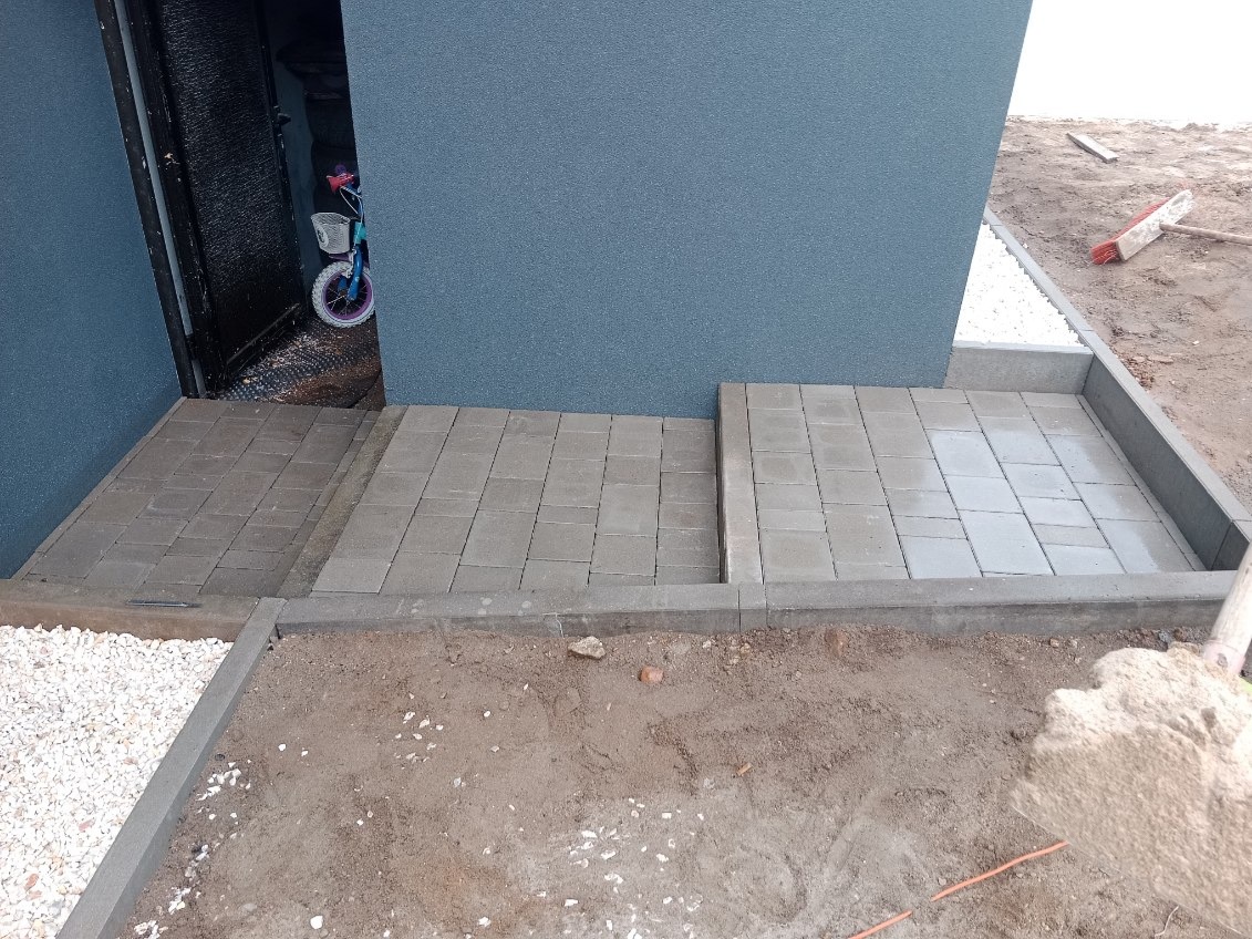 Ułożona szara kostka brukowa tworząca ścieżkę i mały taras przy budynku z niebieską elewacją, w tle widoczny dziecięcy rower.