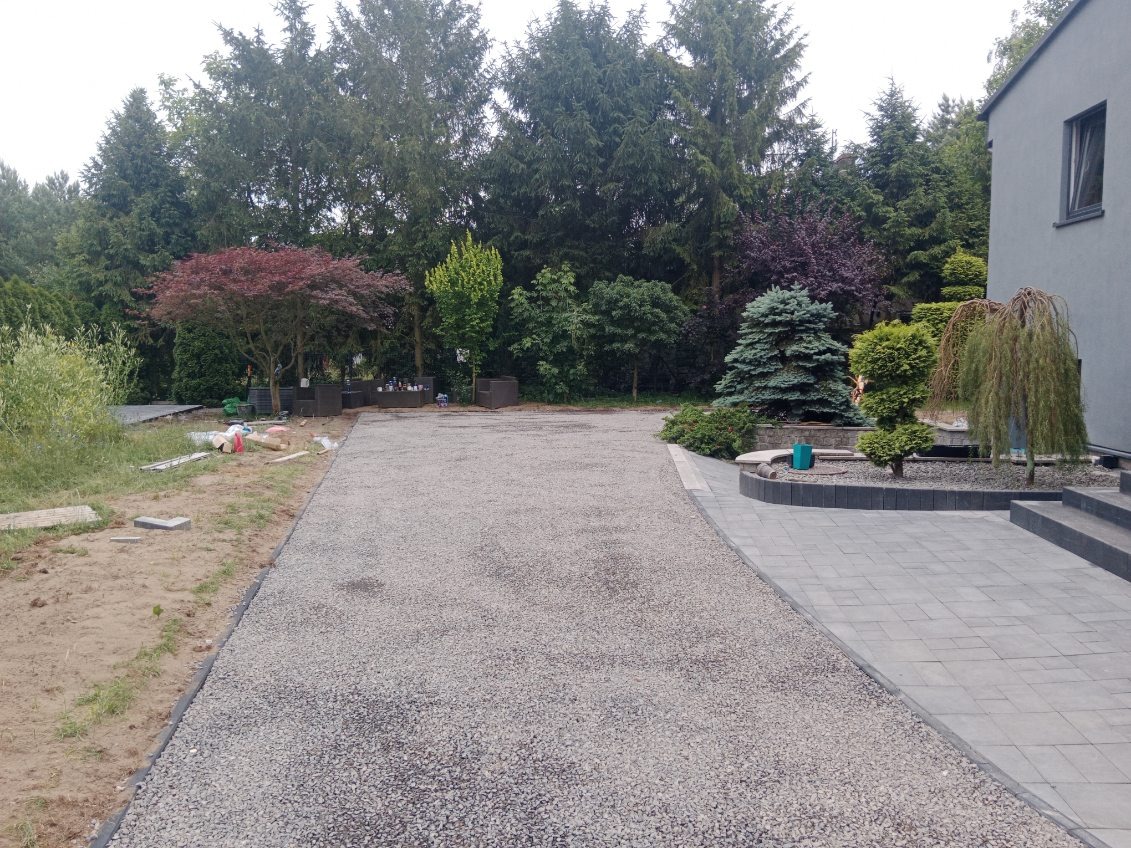 Szeroki podjazd z szarego kruszywa, obrzeża wykończone czarną gumą, z boku ścieżka z szarej kostki brukowej, w tle drzewa i krzewy.