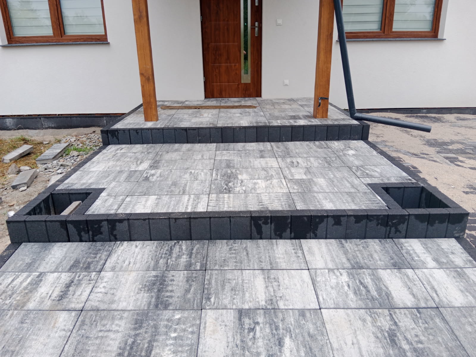 Schody wejściowe wyłożone szarą kostką brukową z czarnymi obrzeżami, widoczne drewniane słupy zadaszenia i fragment elewacji budynku.