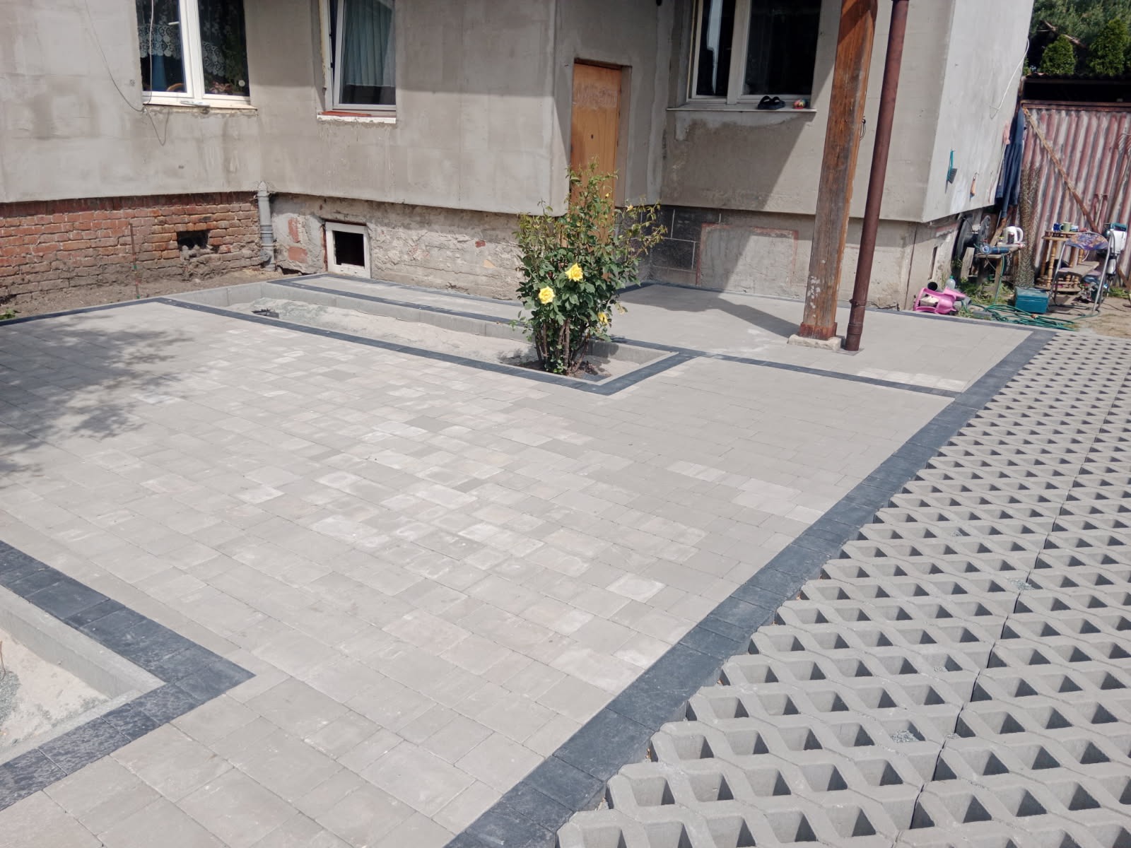 Szara kostka brukowa ułożona na posesji przed domem, z obramowaniem z ciemniejszej kostki i miejscem na roślinę, z boku ażurowa kostka trawnikowa.