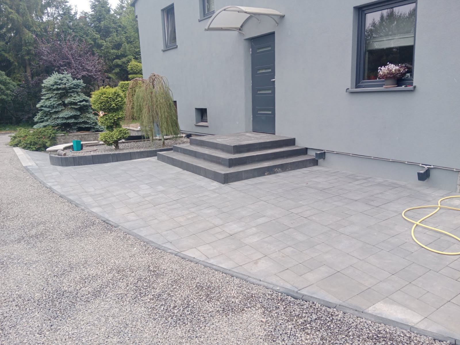 Szara kostka brukowa ułożona przed domem z szarą elewacją, widoczne schody z kostki, obrzeża, rabata z iglakami oraz fragment węża ogrodowego.