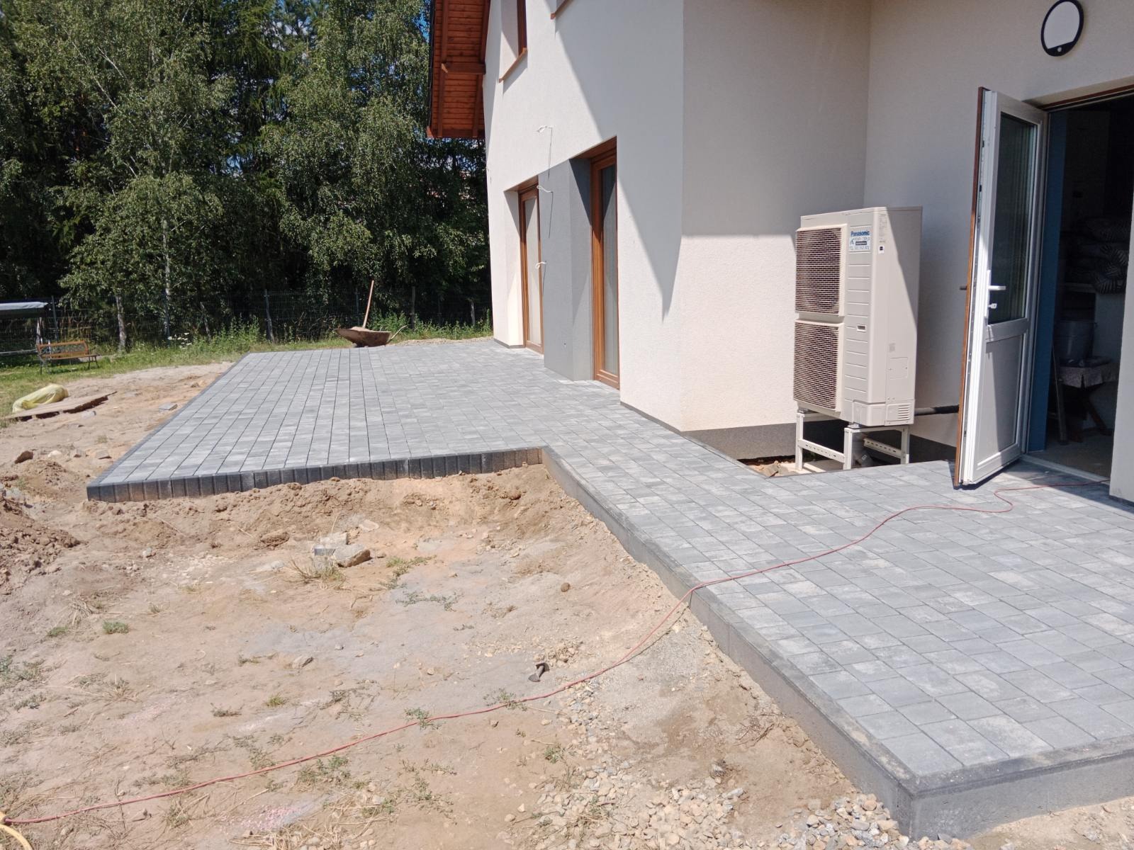 Szara kostka brukowa ułożona wokół domu z widoczną jednostką zewnętrzną klimatyzacji, ziemia przygotowana pod dalsze prace brukarskie, taczka z narzędziami w tle.