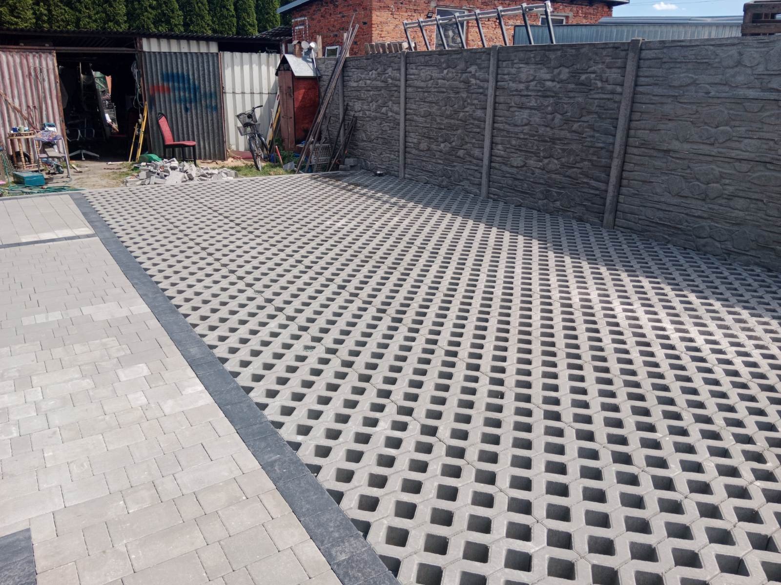 Ułożona szara ażurowa kostka brukowa na podjeździe, obrzeża z ciemnej kostki, tło stanowi betonowe ogrodzenie z fakturą kamienia i blaszany garaż.