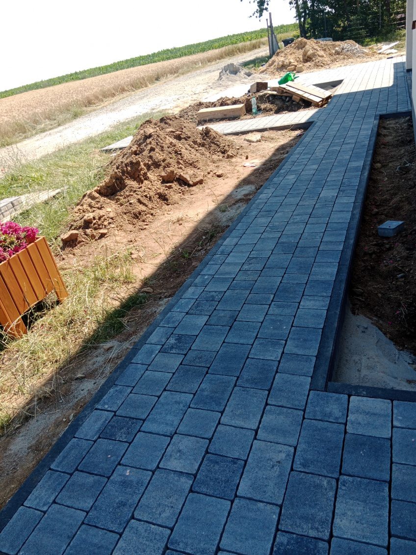 Ułożona ścieżka z ciemnoszarej kostki brukowej prowadząca wzdłuż budynku, z widoczną stertą ziemi i polami w tle, oraz donicą z kwiatami po lewej stronie.