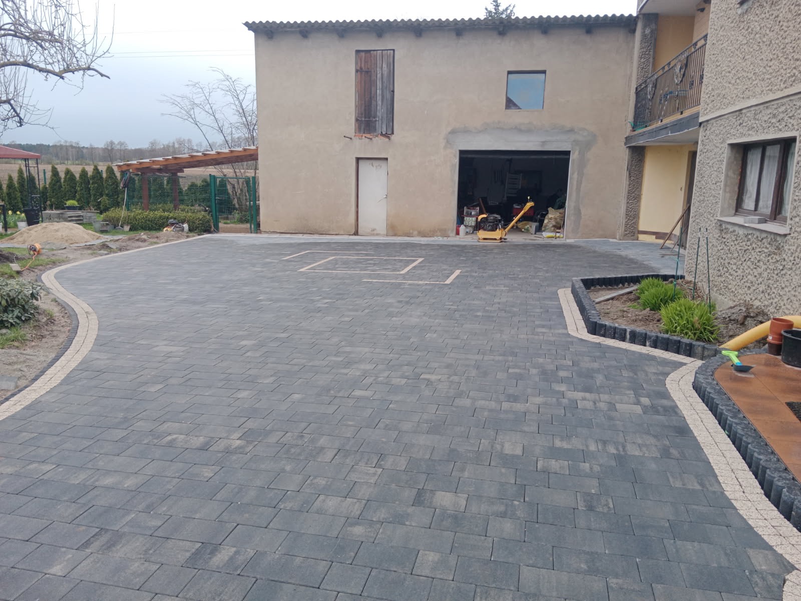 Wykończony podjazd z szarej kostki brukowej z jasną obwódką, widoczny zagęszczarka w tle, zarysowane kwadraty na kostce, obrzeża rabatki z roślinami.