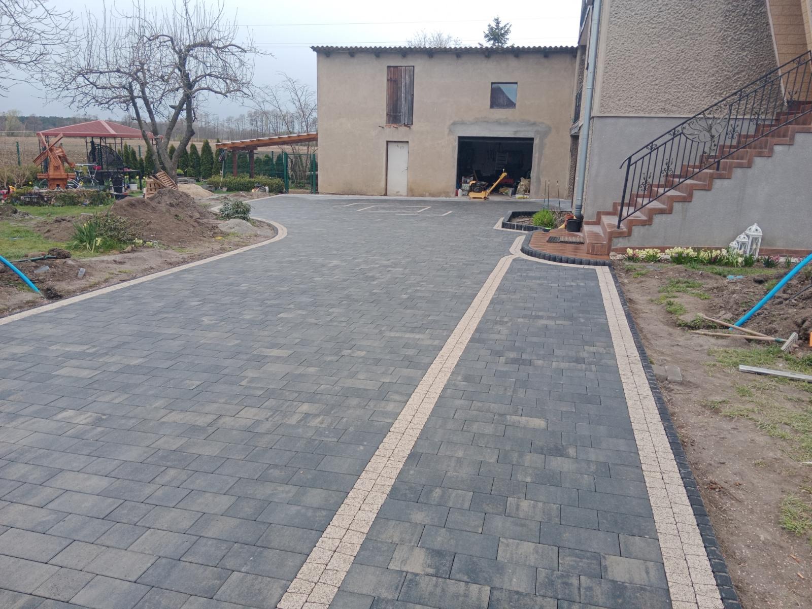 Ułożona kostka brukowa w Żmigrodzie, z jasnymi pasami oddzielającymi, prowadząca do garażu i schodów zewnętrznych, z widoczną ubijarką w tle.