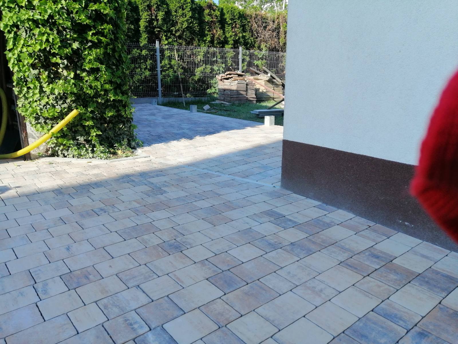 Ułożona kostka brukowa o nieregularnym wzorze i odcieniach beżu, szarości i błękitu, z widocznym fragmentem ściany budynku z cokolem oraz zielenią pnącą się po ścianie, w tle widoczne ogrodzenie...