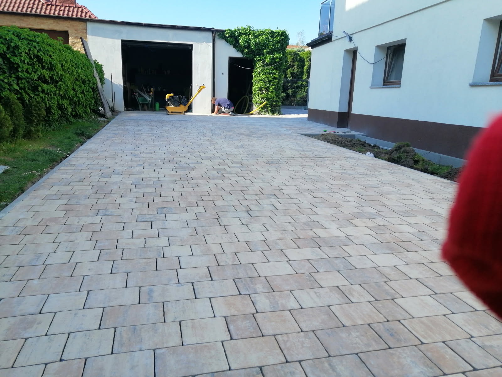 Ułożona beżowo-szara kostka brukowa na podjeździe, widoczna ubijarka w tle, budynek z garażem i fragment trawnika.