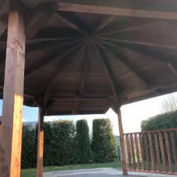 Drewniana pergola z widoczną konstrukcją dachu i częścią ogrodzenia, otoczona zielenią.