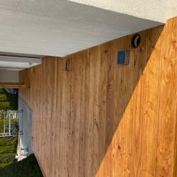 kaspol - Wykończona drewniana elewacja tarasu z widoczną strukturą drewna, zamontowany element oświetleniowy i kratka wentylacyjna, w tle widoczny ogród i drabina