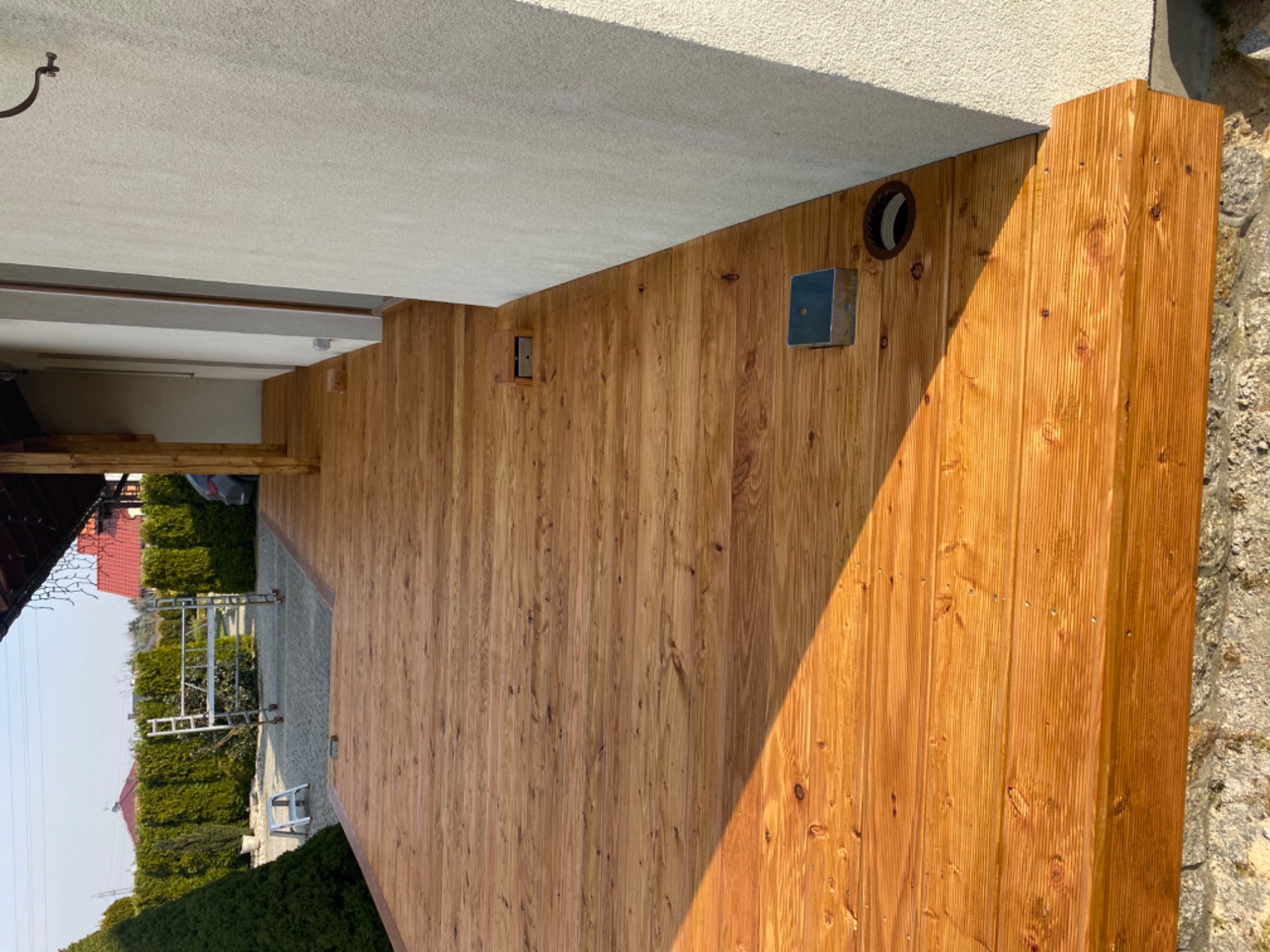 Wykończona drewniana elewacja tarasu z widoczną strukturą drewna, zamontowany element oświetleniowy i kratka wentylacyjna, w tle widoczny ogród i drabina