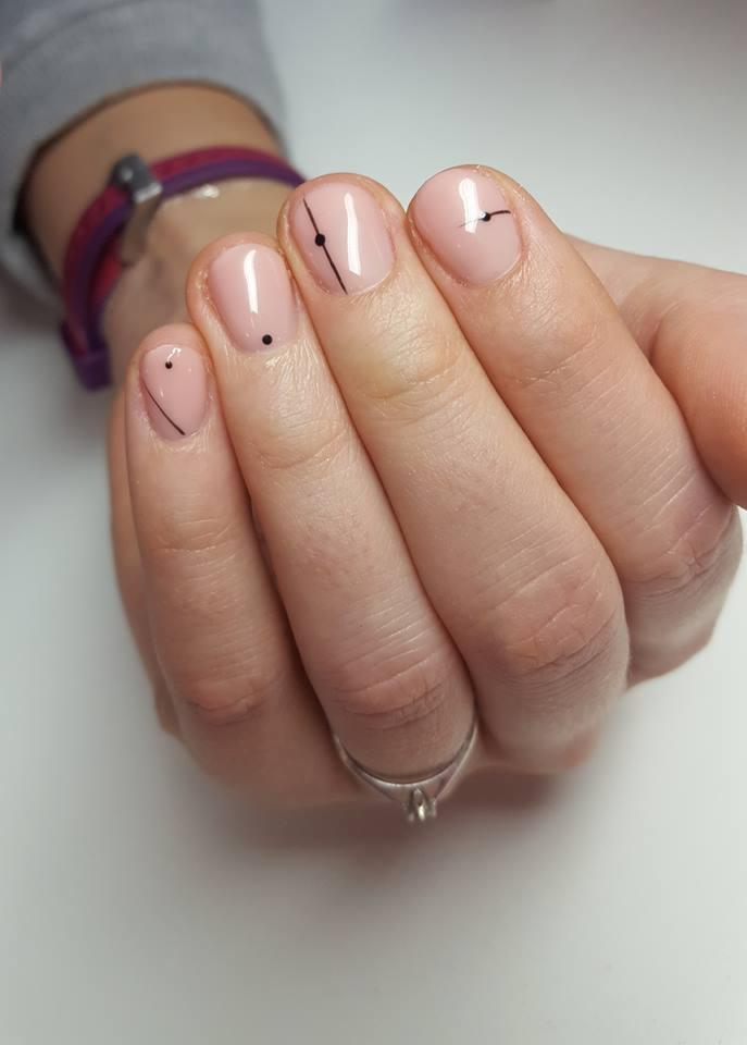 Dłoń z delikatnym, różowym manicure z minimalistycznym czarnym wzorem: kropki i kreski na każdym paznokciu. Na palcu serdecznym srebrny pierścionek z oczkiem. W tle fragment ubrania z bransoletką.