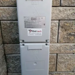 Dwa połączone szare panele sterowania DEA na kamiennej ścianie zewnętrznej, z naklejką Fiber.Tech i informacjami technicznymi.