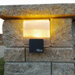 Kwadratowa lampa zewnętrzna z logo 'Nice' zamontowana na kamiennym słupku ogrodzenia, oświetlona ciepłym światłem.
