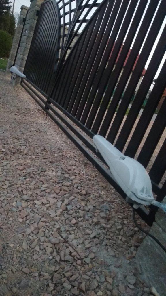 Czarna brama przesuwna z dwoma zamontowanymi napędami automatycznymi w szarych obudowach, widok z boku na żwirowe podłoże.