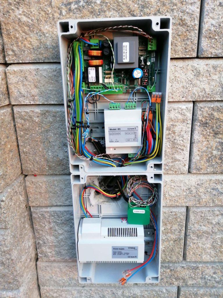 Otwarta obudowa domofonu z widocznymi podzespołami elektronicznymi, kablami i zasilaczem zamontowana na ścianie z kamienia dekoracyjnego.