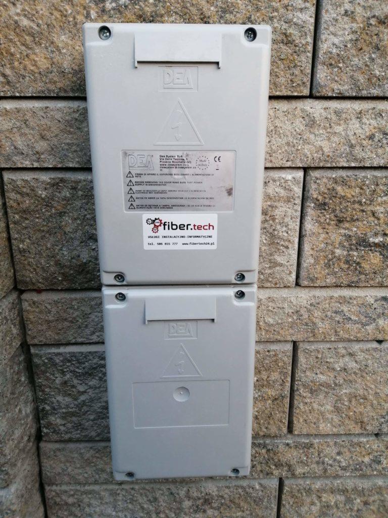 Dwa połączone szare panele sterowania DEA na kamiennej ścianie zewnętrznej, z naklejką Fiber.Tech i informacjami technicznymi.